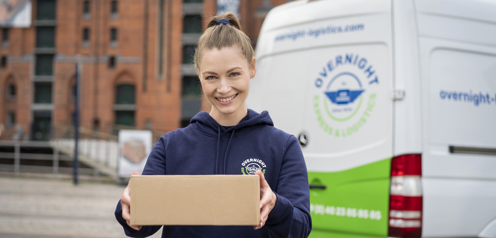 Eine lächelnde Frau hält ein Paket in den Händen, im Hintergrund ist ein Lieferwagen mit der Aufschrift „Overnight Express & Logistics“ zu sehen.