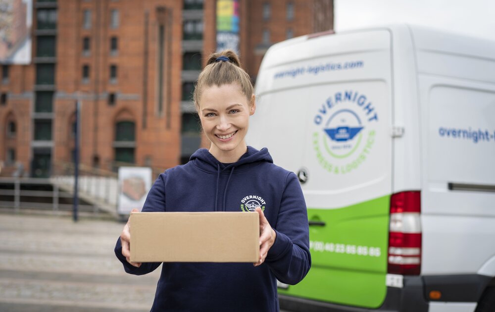 Eine lächelnde Frau hält ein Paket in den Händen, im Hintergrund ist ein Lieferwagen mit der Aufschrift „Overnight Express & Logistics“ zu sehen.