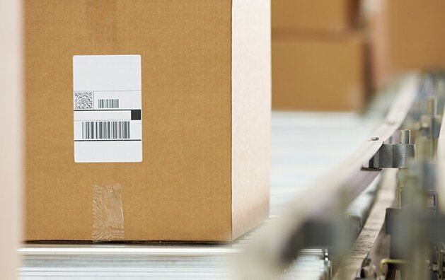 Ein Karton mit einem Etikett steht auf einem Förderband, umgeben von weiteren Kisten im Hintergrund.