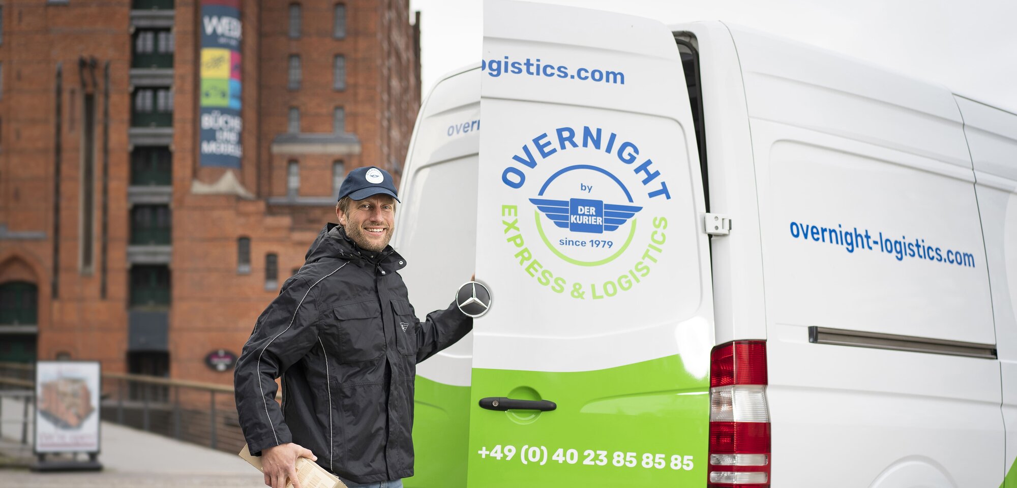 Ein Lieferfahrer steht neben einem weißen Lieferwagen mit der Aufschrift „Overnight Express & Logistics“ und hält ein Paket in der Hand.