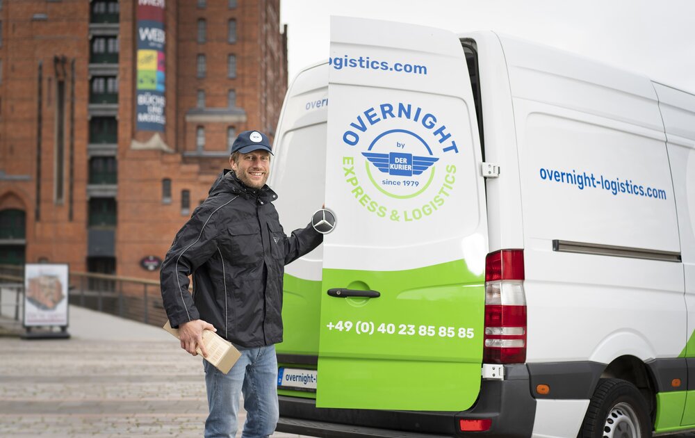 Ein Lieferfahrer steht neben einem weißen Lieferwagen mit der Aufschrift „Overnight Express & Logistics“ und hält ein Paket in der Hand.