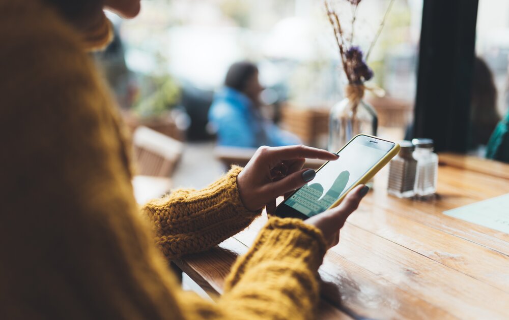 Eine Person in einem gelben Pullover hält ein Smartphone und tippt darauf, während sie an einem Holztisch in einem Café sitzt.
