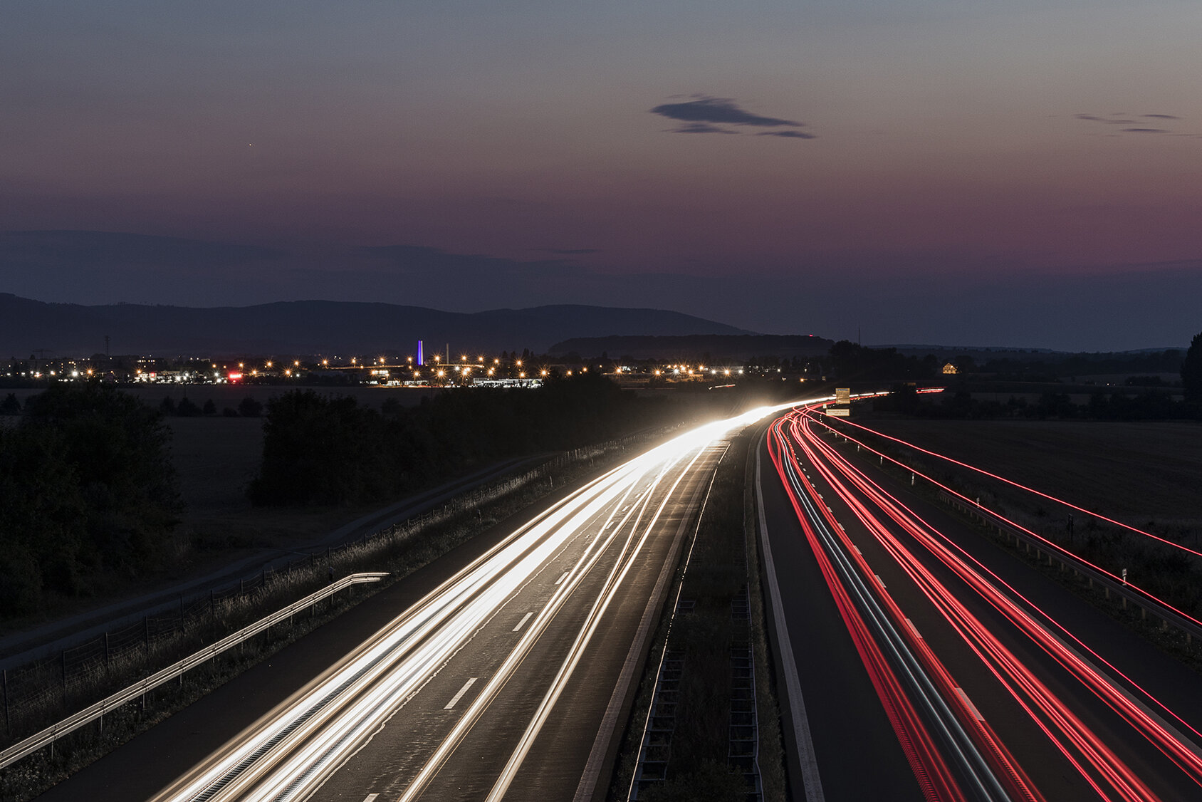 Lichtspuren von Fahrzeugen auf einer Autobahn bei Dämmerung, mit einer städtischen Umgebung im Hintergrund und Bergen in der Ferne.