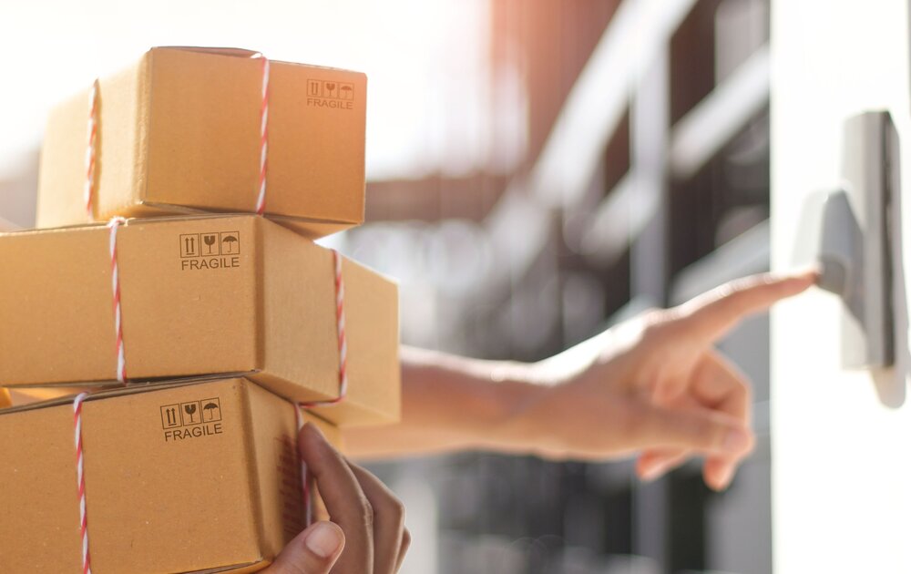 Eine Person hält mehrere Pakete und drückt an einer Klingel. Die Pakete sind mit einem Bindfaden umwickelt und tragen den Aufdruck „Fragile“.