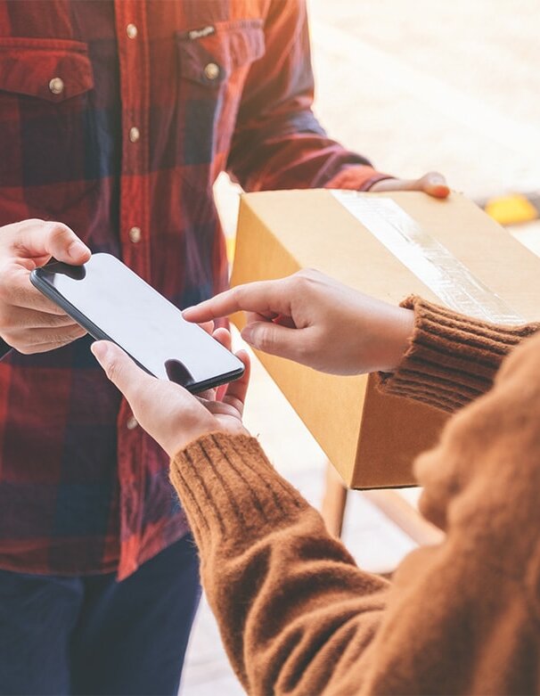 Eine Person übergibt ein Packet an eine andere Person, während sie auf ein Smartphone zeigt. Beide stehen an einer Haustür.