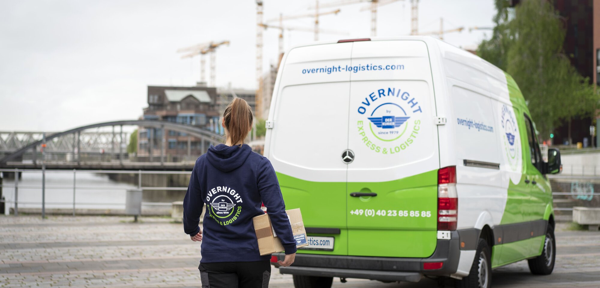 Eine Person trägt ein Paket und geht auf einen Lieferwagen mit der Aufschrift „Overnight Express & Logistics“ zu. Im Hintergrund ist eine Brücke und eine Stadtansicht sichtbar.
