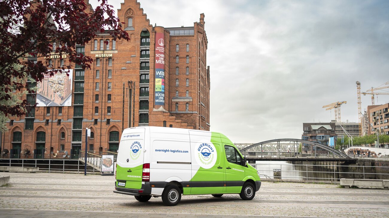 Ein grüner Lieferwagen steht auf einem Pflasterweg vor einem historischen Backsteingebäude, das mit Werbetafeln dekoriert ist. Im Hintergrund ist eine Brücke sichtbar.