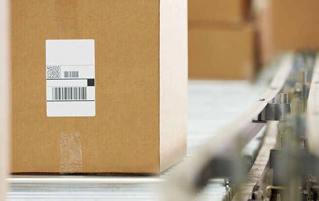 Ein Karton mit einem Barcode steht auf einem Förderband in einem Lager. Im Hintergrund sind weitere Kartons sichtbar.