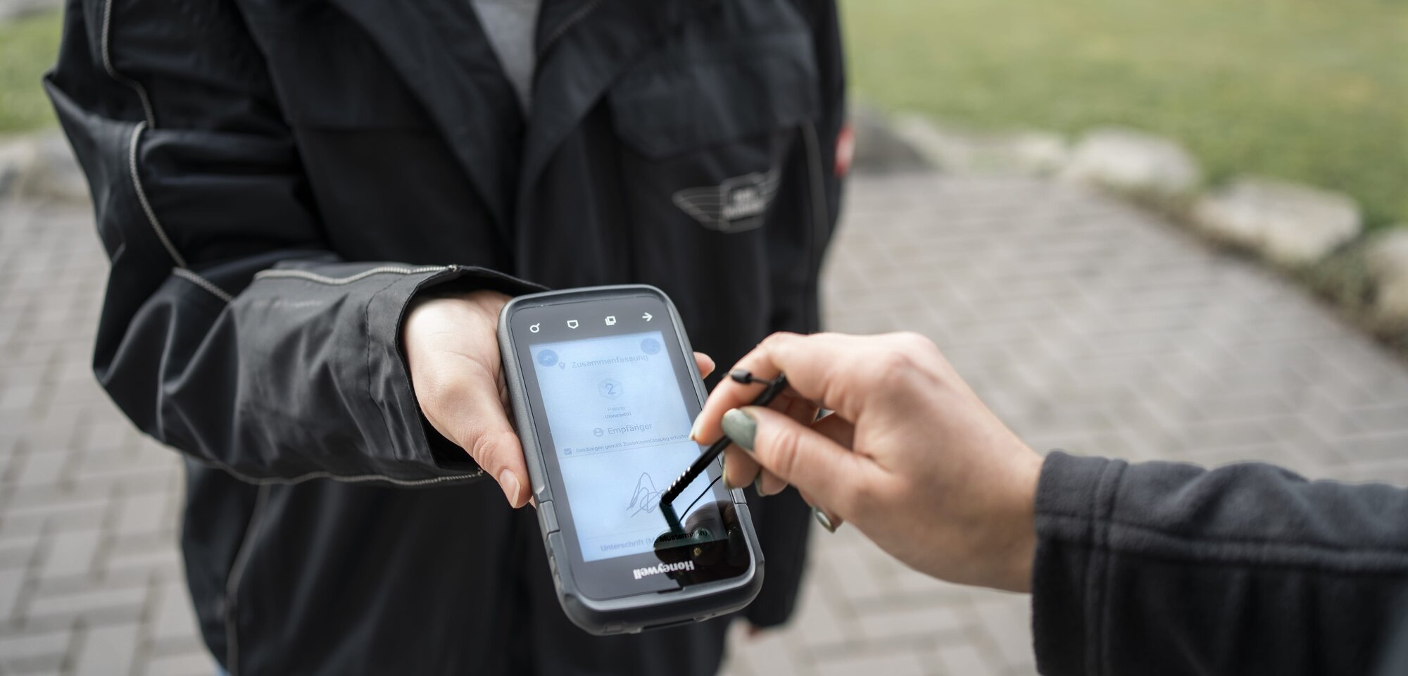 Eine Person hält ein mobiles Gerät, während eine andere Person mit einem Stylus darauf unterschreibt. Das Gerät zeigt eine digitale Benutzeroberfläche.
