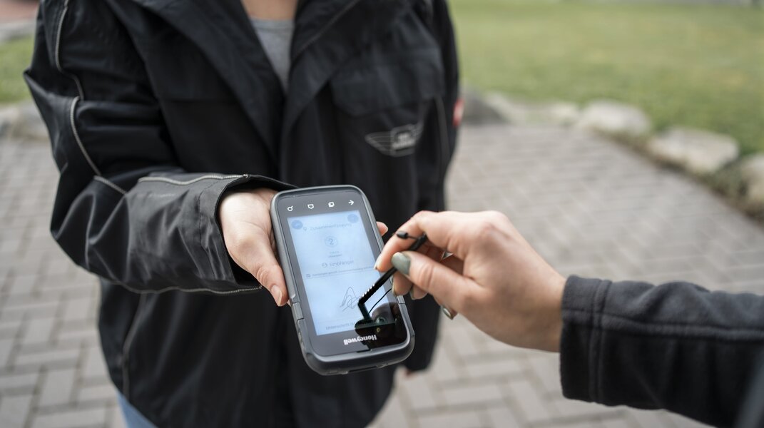 Eine Person hält ein mobiles Gerät, während eine andere Person mit einem Stylus darauf unterschreibt. Das Gerät zeigt eine digitale Benutzeroberfläche.