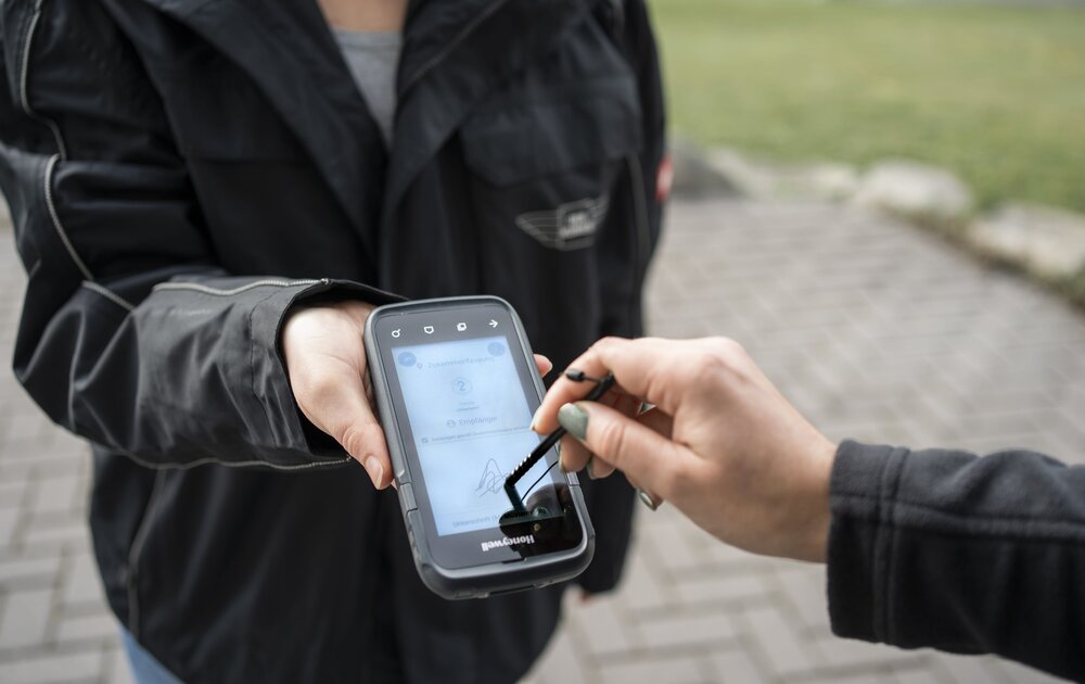 Eine Person hält ein mobiles Gerät, während eine andere Person mit einem Stylus darauf unterschreibt. Das Gerät zeigt eine digitale Benutzeroberfläche.