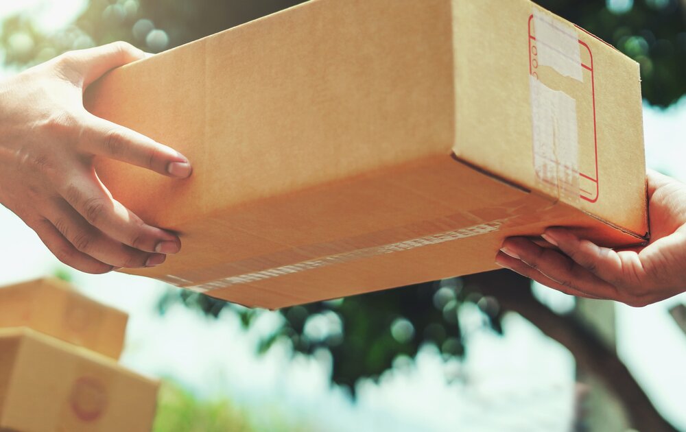 Ein Zusteller übergibt ein Paket an eine andere Person. Beide Hände sind sichtbar, während im Hintergrund Bäume zu sehen sind.