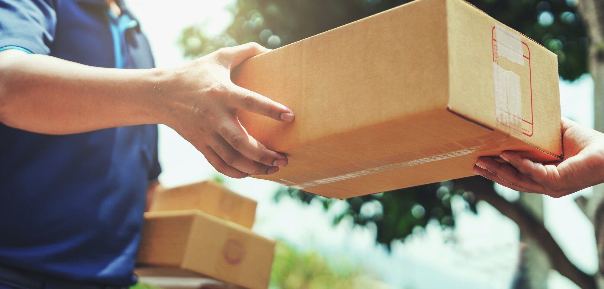 Ein Zusteller übergibt ein Paket an eine andere Person. Beide Hände sind sichtbar, während im Hintergrund Bäume zu sehen sind.