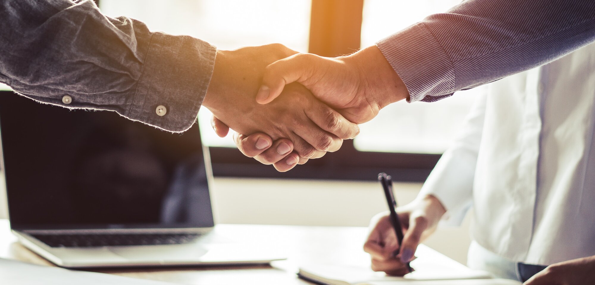 Zwei Personen schütteln sich die Hände, während ein Laptop und Notizen auf dem Tisch liegen. Das Bild symbolisiert eine geschäftliche Vereinbarung oder Partnerschaft.