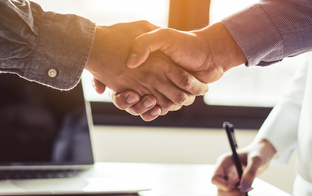Zwei Personen schütteln sich die Hände, während ein Laptop und Notizen auf dem Tisch liegen. Das Bild symbolisiert eine geschäftliche Vereinbarung oder Partnerschaft.