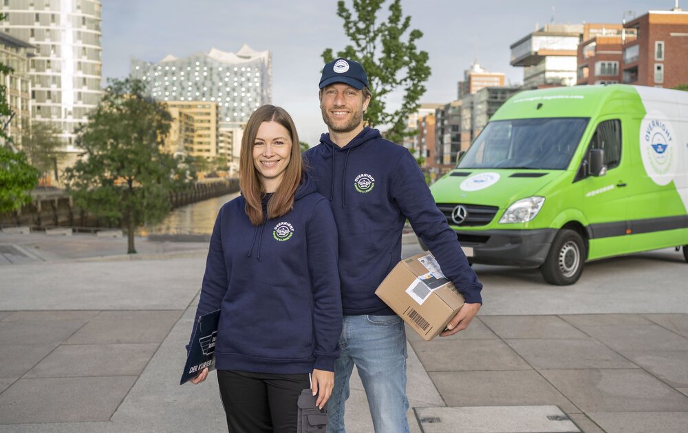 Ein Mann und eine Frau stehen lächelnd vor einem Lieferwagen, beide in blauen Kapuzenpullis. Der Mann hält ein Paket in der Hand.