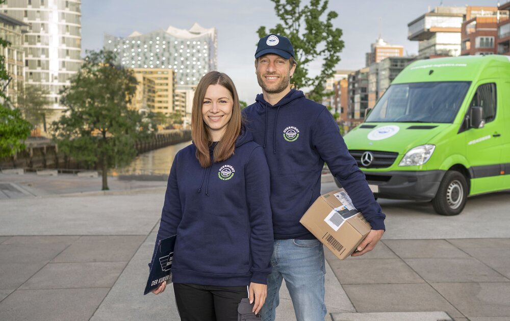 Ein Mann und eine Frau stehen lächelnd vor einem Lieferwagen, beide in blauen Kapuzenpullis. Der Mann hält ein Paket in der Hand.