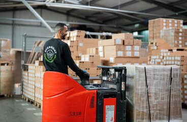 Mitarbeiter von Overnight Express & Logistics bewegt mit einem Elektro-Hubwagen eine folierte Palette in einer Lagerhalle mit gestapelten Kartons