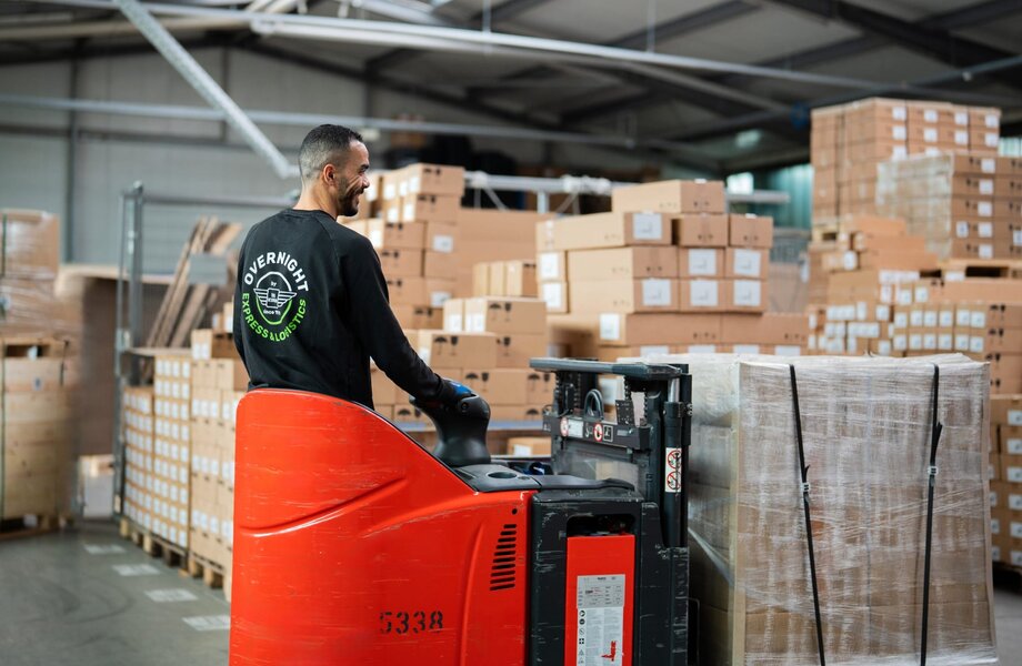 Overnight Express & Logistics: Warenumschlag im Lager Mitarbeiter von Overnight Express & Logistics bewegt mit einem Elektro-Hubwagen eine folierte Palette in einer Lagerhalle mit gestapelten Kartons