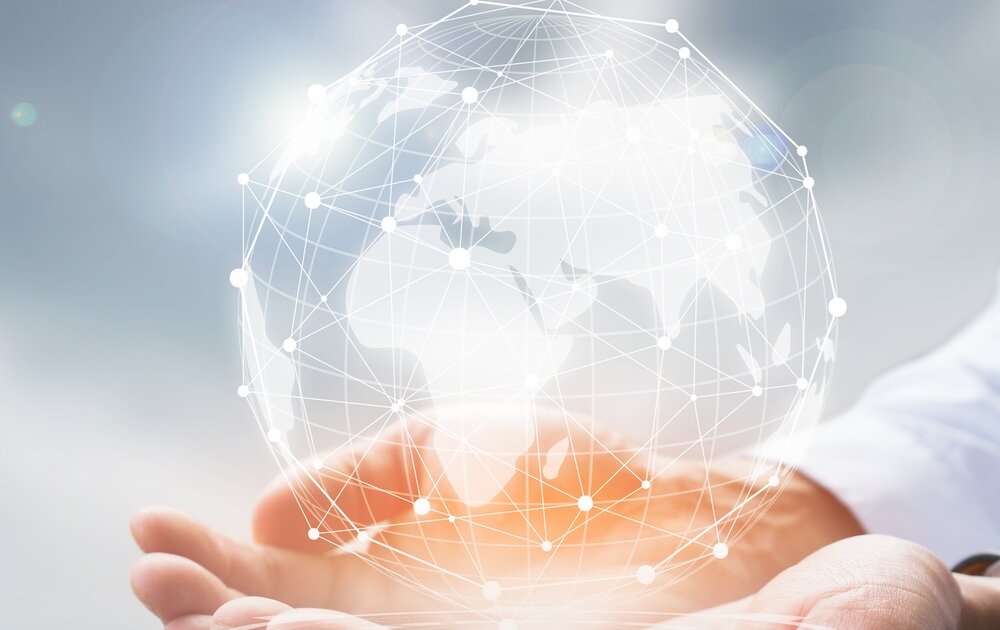 Eine Hand hält eine transparente, vernetzte Weltkugel, die globale Vernetzung und Technologie symbolisiert, vor einem sanften, verschwommenen Hintergrund.