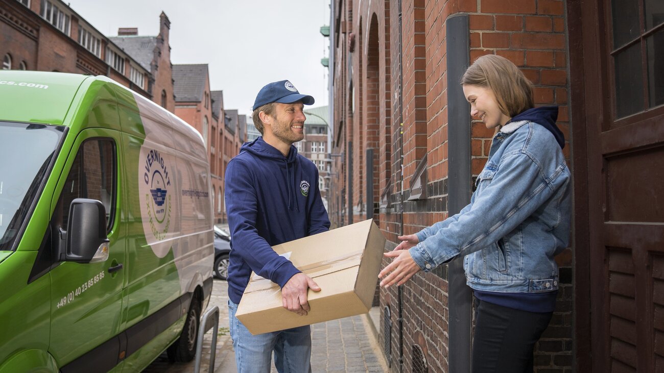 Ein "Overnight Express & Logistics" Kurier übergibt ein Paket an einen Kunden. Im Hintergrund ist der Transporter von "Overnight Express & Logistics" zu sehen.