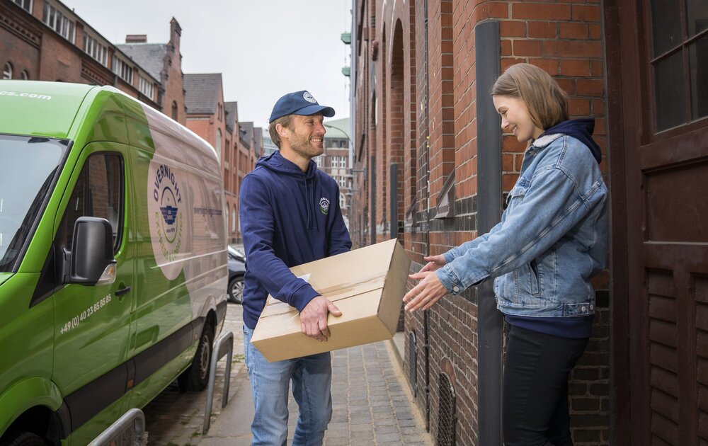Ein "Overnight Express & Logistics" Kurier übergibt ein Paket an einen Kunden. Im Hintergrund ist der Transporter von "Overnight Express & Logistics" zu sehen.