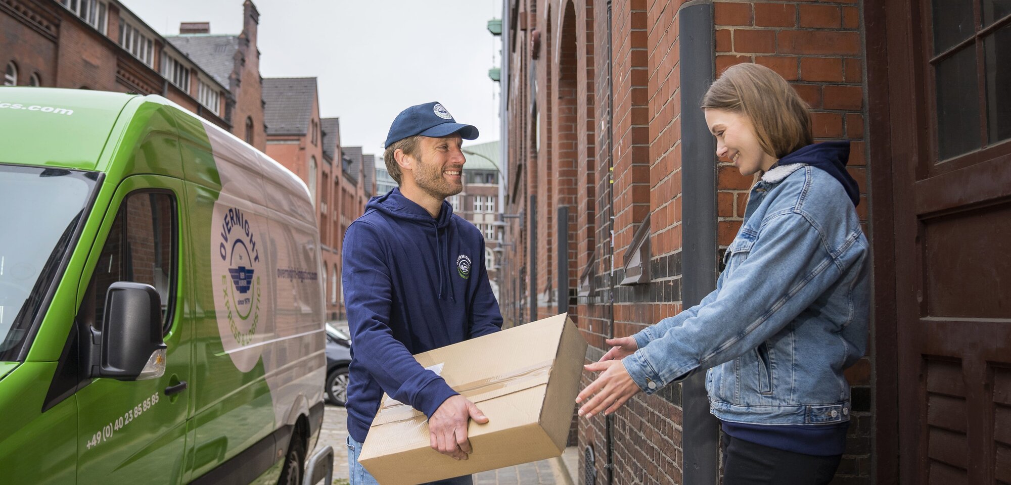 Ein "Overnight Express & Logistics" Kurier übergibt ein Paket an einen Kunden. Im Hintergrund ist der Transporter von "Overnight Express & Logistics" zu sehen.