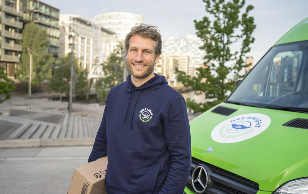 Ein lächelnder Mann mit einem Karton steht vor einem grünen Lieferfahrzeug. Im Hintergrund sind moderne Gebäude und Bäume zu sehen.