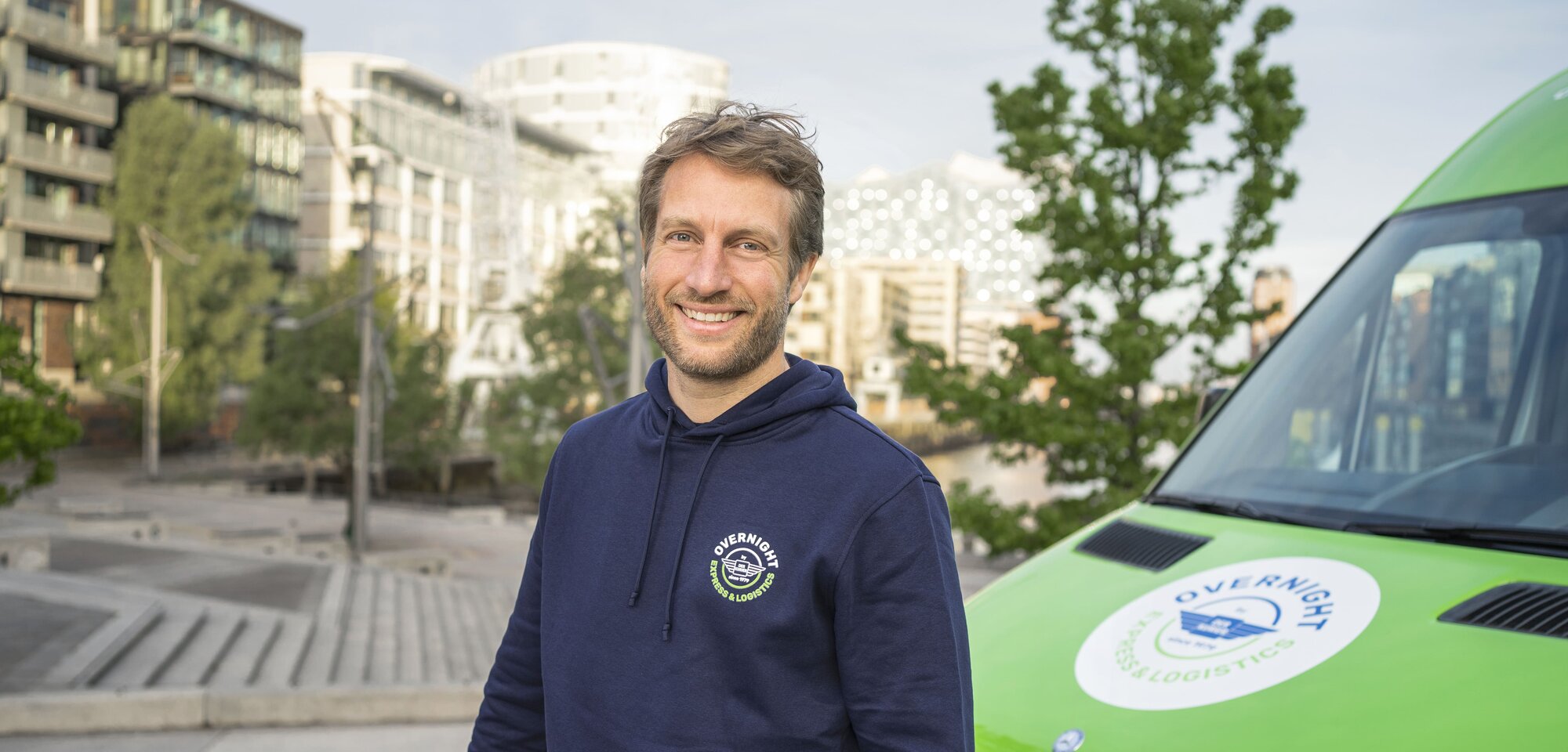 Ein lächelnder Mann mit einem Karton steht vor einem grünen Lieferfahrzeug. Im Hintergrund sind moderne Gebäude und Bäume zu sehen.