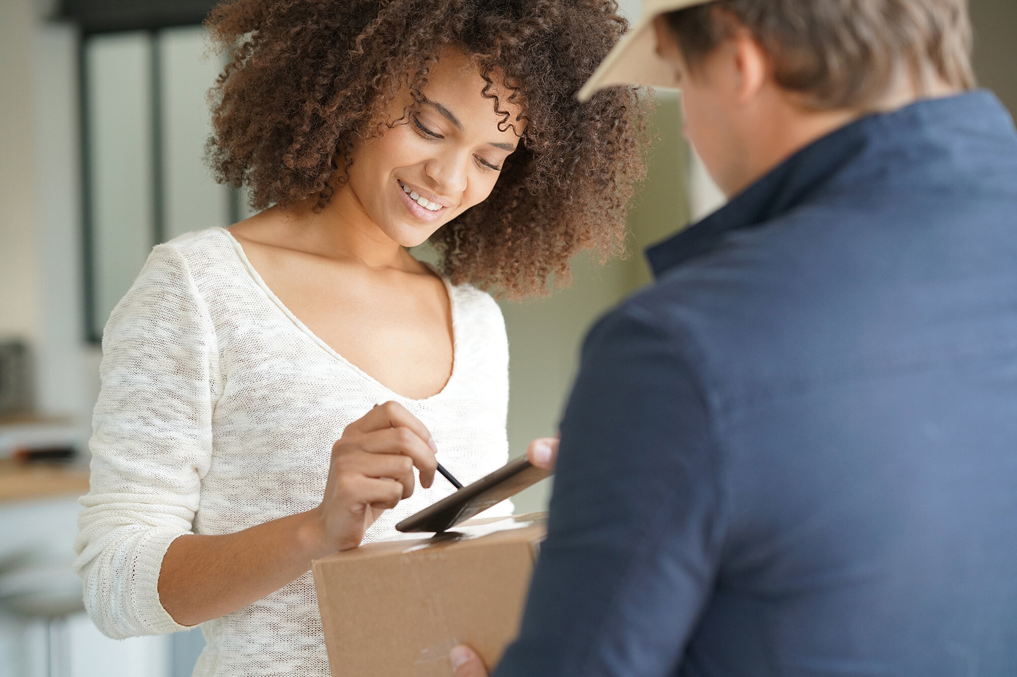 Eine junge Frau mit lockigem Haar signiert auf einem Tablet, während sie ein Paket entgegenimmt. Ihre Miene strahlt Freude aus.
