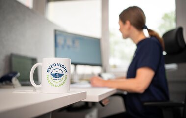 Tasse mit Overnight Express & Logistics Logo auf einem Schreibtisch, im Hintergrund eine Mitarbeiterin am Büroarbeitsplatz